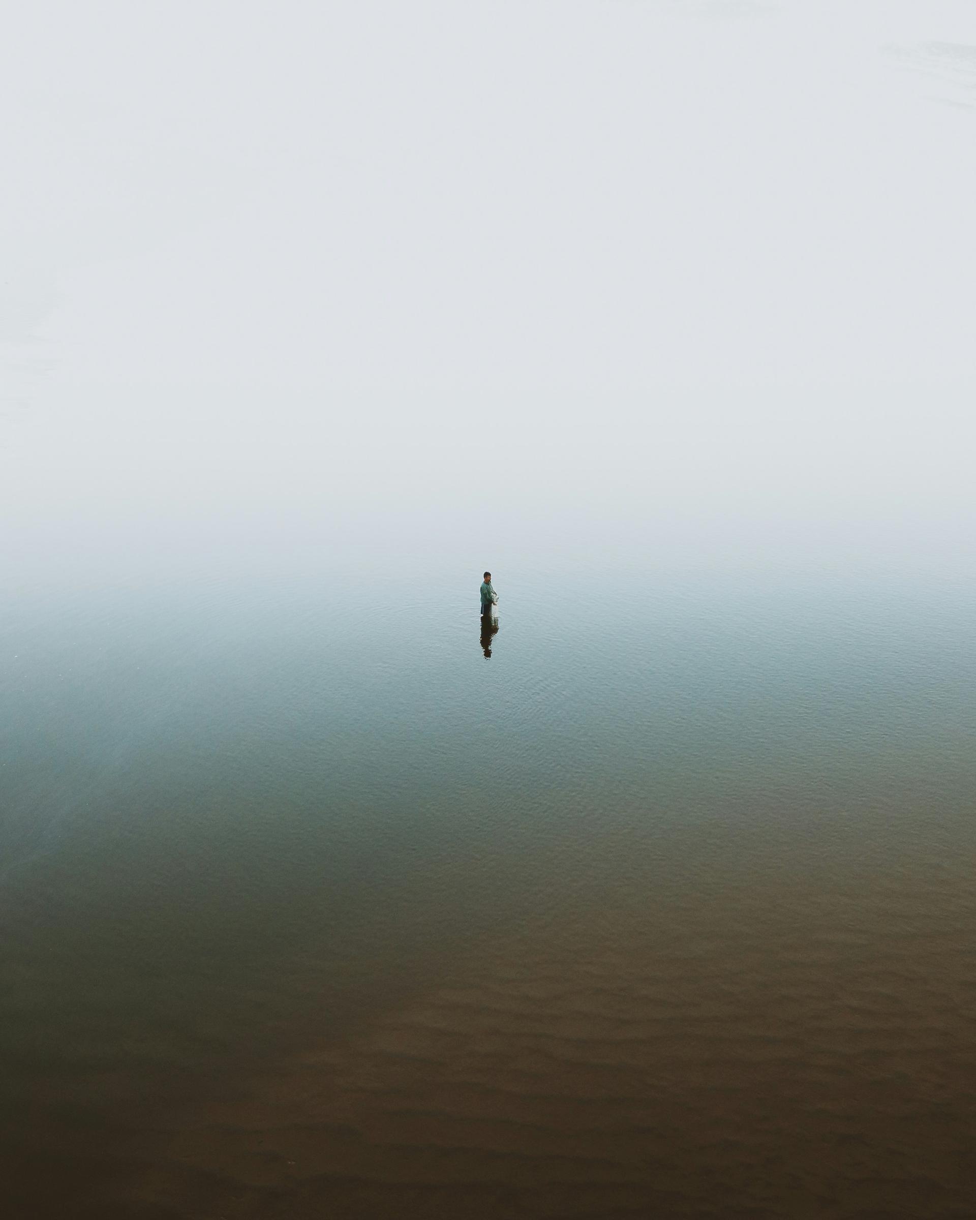 A lone fisherman stands waist-deep in a misty body of water during daytime, surrounded by fog, creating a serene and isolated atmosphere